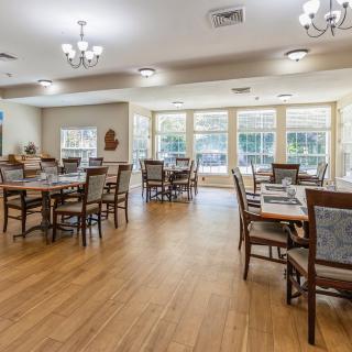 A spacious dining room with multiple tables and chairs, featuring wooden flooring and large windows.