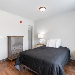 A minimalist bedroom with a bed, dresser, and lamp on a wooden floor.