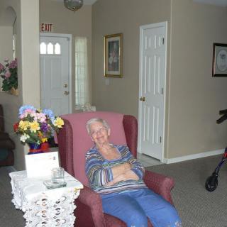 An elderly woman sitting comfortably in an armchair in a cozy living room with flowers and a walker nearby.
