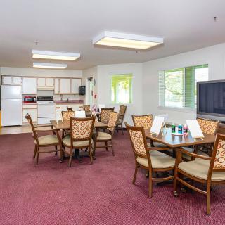 A communal dining area with several tables and chairs, a kitchen in the background, and a television in the corner.