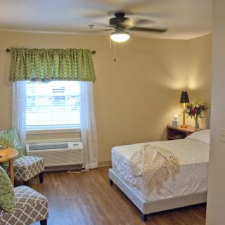 A cozy bedroom with a neatly made bed, two chairs, a small table, and a window adorned with green curtains.
