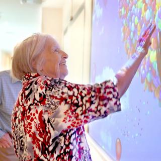 An elderly woman engaging with interactive art on a digital screen, smiling as she touches the colorful display.