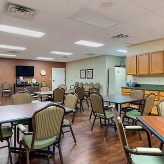 A spacious community room with multiple tables and chairs arranged for gatherings, featuring a kitchenette in the corner.