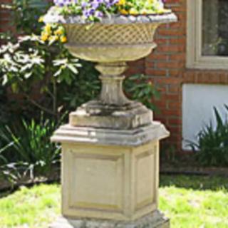 A stone urn planter filled with colorful flowers on a pedestal in a garden setting.