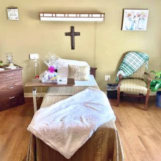 A cozy hospital room featuring a bed with a blanket, a small table with flowers, and a chair for visitors.