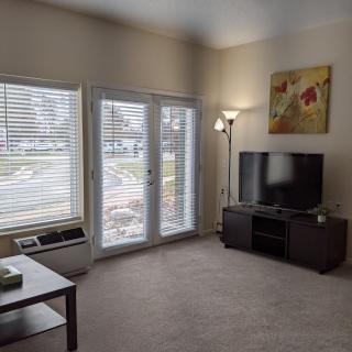 A cozy living room with a television, carpet, and large windows with blinds.