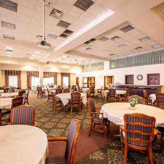 A spacious dining room with round tables and upholstered chairs, featuring warm lighting and carpeted flooring.