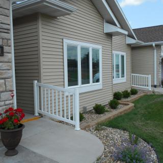 A house exterior with beige siding, white-trimmed windows, and a small porch with railing surrounded by neatly landscaped plants.