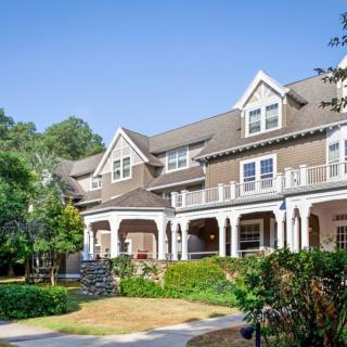 A large, multi-story residential house with a spacious porch and well-maintained landscaping under a clear blue sky.