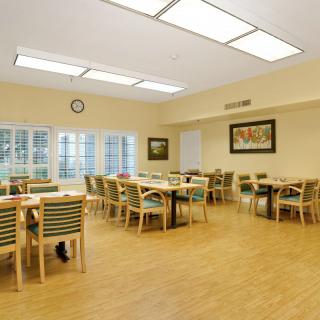 A spacious dining room with multiple tables and chairs, illuminated by overhead lighting and natural light from large windows.