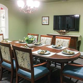 A formal dining room with a large table set for eight, illuminated by a chandelier and featuring a wall-mounted television.