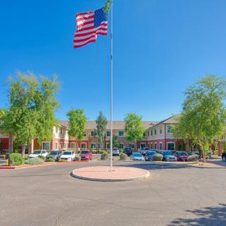 A view of a building complex with an American flag flying in front, surrounded by parked cars and trees under a clear blue sky.