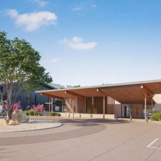 A modern building with a sleek wooden entrance, surrounded by landscaping and trees against a clear sky.