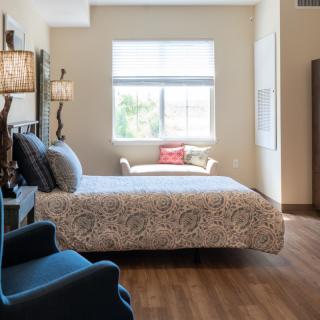 A cozy bedroom with ample natural light, featuring a bed, chair, and wooden flooring.