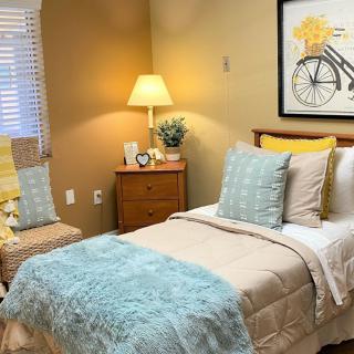 A cozy bedroom with a neatly made bed, featuring soft blue and beige bedding, decorative pillows, and warm lighting from a bedside lamp.