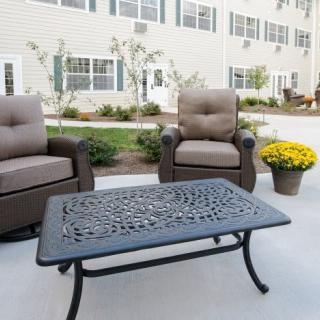 A patio seating area with two cushioned chairs and a decorative metal table, complemented by a pot of yellow mums in front of a building.