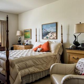 A cozy bedroom with a neatly made bed, adorned with orange and white pillows, flanked by wooden nightstands and lamps. Art hangs on the walls, adding a touch of elegance to the room.