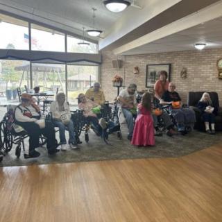A group of seniors gathered in a nursing home common area, some seated in wheelchairs.