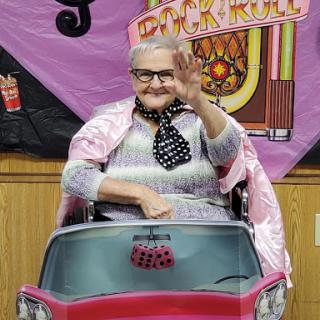 An elderly woman in a pink cape greets warmly while sitting in a toy car, embodying joy and vintage charm.