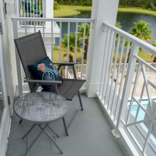 A cozy balcony setup with a chair, cushion, and table overlooking a lake and palm trees.