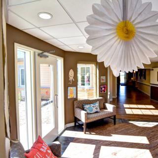 A brightly lit lobby area with decorative elements, including a large daisy hanging from the ceiling and comfortable seating near the entrance.