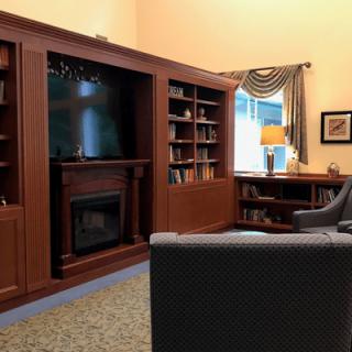 A cozy living room with wooden bookshelves, a fireplace, and a television. It also features a seating area and a desk near the window.