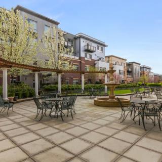 A pleasant outdoor patio with tables and chairs surrounded by apartment buildings, featuring a fountain and a pergola on a sunny day.