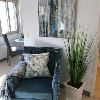 A cozy corner featuring a blue accent chair with a floral pillow and throw, complemented by a tall plant and abstract wall art.