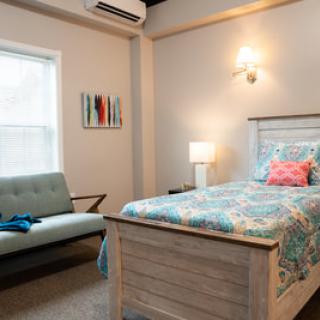 A cozy bedroom with a single bed featuring colorful bedding, a small sofa, and a lamp by the window.