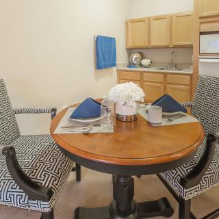 A cozy dining area featuring a wooden round table set for two, with patterned chairs and a minimalist kitchen backdrop.
