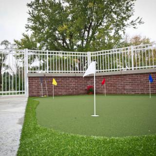 An outdoor putting green with multiple colored flags, set against a brick wall and fence.