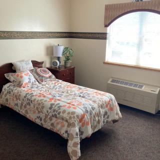 A cozy bedroom with a single bed featuring floral bedding, a wooden nightstand with a lamp, and a large window allowing natural light in.