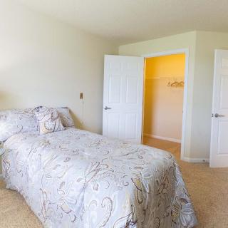 A bright bedroom with a bed and open closet doors, featuring light-colored walls and carpet.