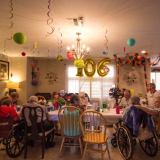 A festive celebration for a 106th birthday with friends and family gathered around a table, surrounded by colorful decorations and balloons indoors.