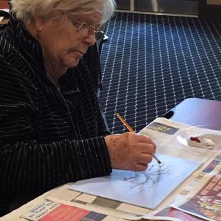 An elderly woman is drawing with a pencil on paper at a table covered with newspapers.