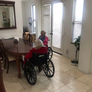 Two elderly individuals in wheelchairs sitting at a dining table in a bright, tiled-floor room.
