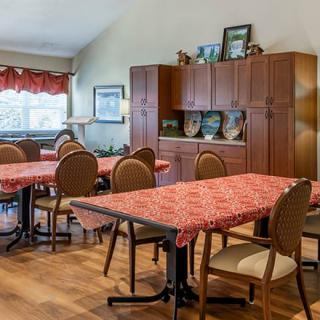 A cozy dining area with tables covered in red-patterned tablecloths, surrounded by wooden chairs and cabinets adorned with decorations.