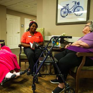 A caretaker sitting with two elderly women, one in a wheelchair and another with a walker, engaged in conversation.