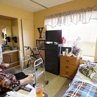 An elderly person sitting in a wheelchair in a nursing home room, with a walker nearby and personal belongings on the dresser.
