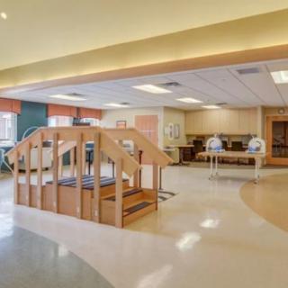 A spacious rehabilitation center therapy room featuring exercise equipment, a wooden staircase, and parallel bars.