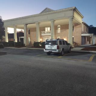 A view of a well-lit building with columns, featuring a parked vehicle in the foreground during twilight.