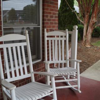 Two white rocking chairs on a porch outside a brick house.