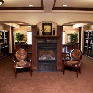 A cozy library interior with a fireplace, flanked by two chairs and surrounded by bookshelves.