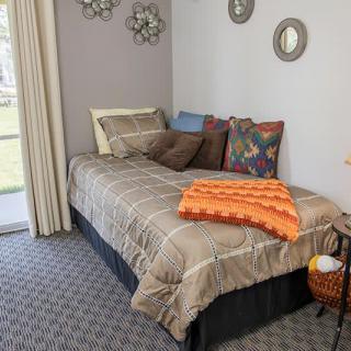 A cozy bedroom with a single bed adorned with colorful pillows and a patterned blanket, featuring wall decor and carpeted flooring.