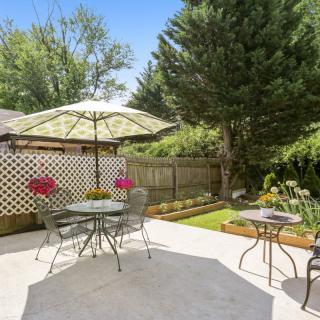A sunny backyard patio featuring outdoor furniture with an umbrella, surrounded by a lush garden and fence.