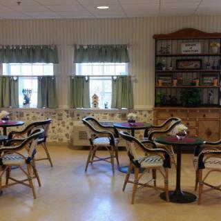 A cozy dining area with decorated tables and chairs, featuring windows with curtains and a wooden cabinet.