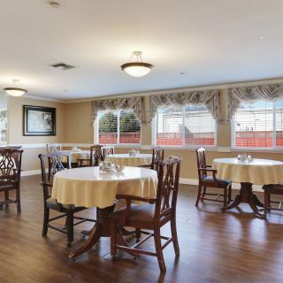 A cozy dining area with neatly arranged tables and chairs, wooden flooring, and large windows dressed with curtains.