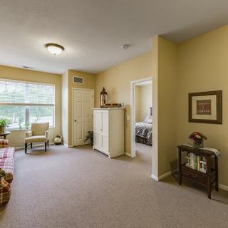A cozy living area with neutral tones, featuring a comfortable armchair, carpet flooring, and ample natural light.