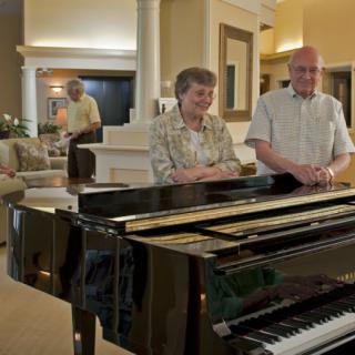 An elderly couple standing beside a grand piano in a well-furnished living room.