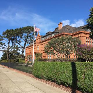 A large brick mansion with a manicured hedge and an American flag flying on a sunny day.